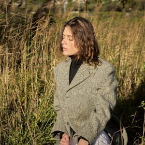 A woman with eyes closed poses in a field, wearing a tweed jacket. Natural sunlight.