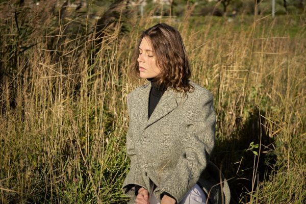 A woman with eyes closed poses in a field, wearing a tweed jacket. Natural sunlight.